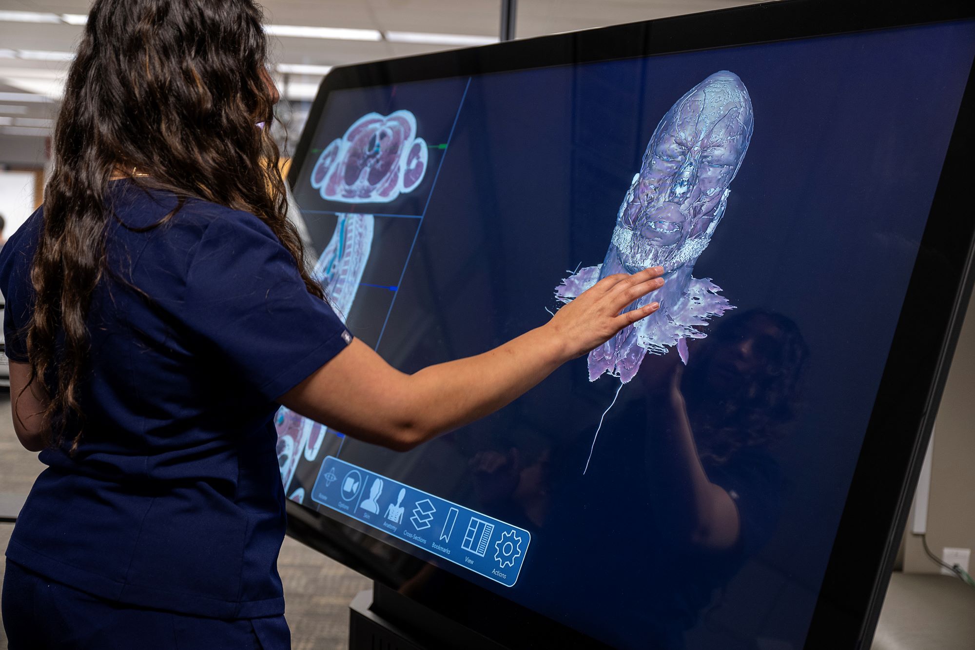 Woman in blue scrubs touching a large monitor touchscreen, interacting with an anatomical human face.