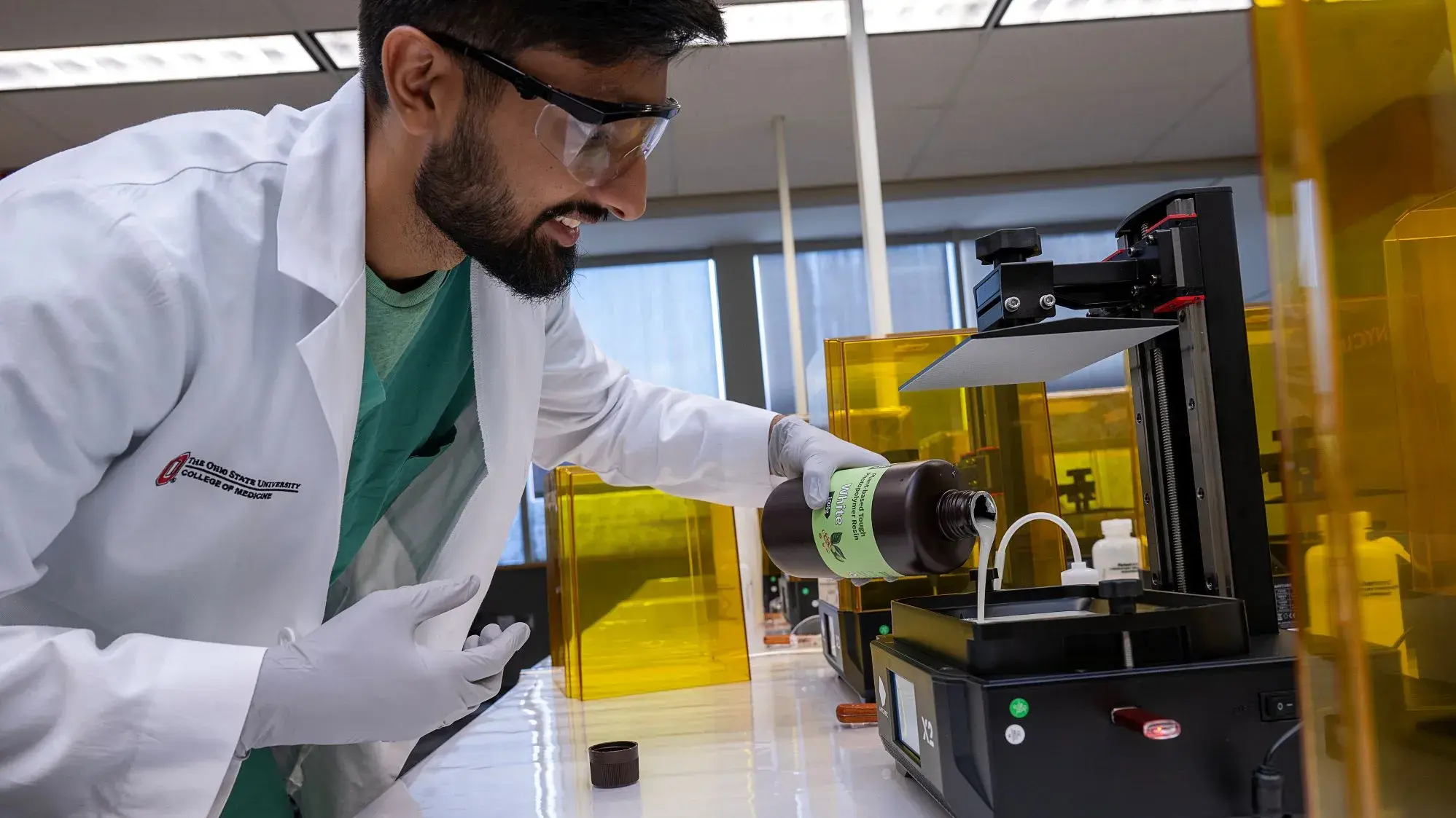 Ohio State student in lab coat and safety equipment pouring resin into a tray of a 3D printer device.