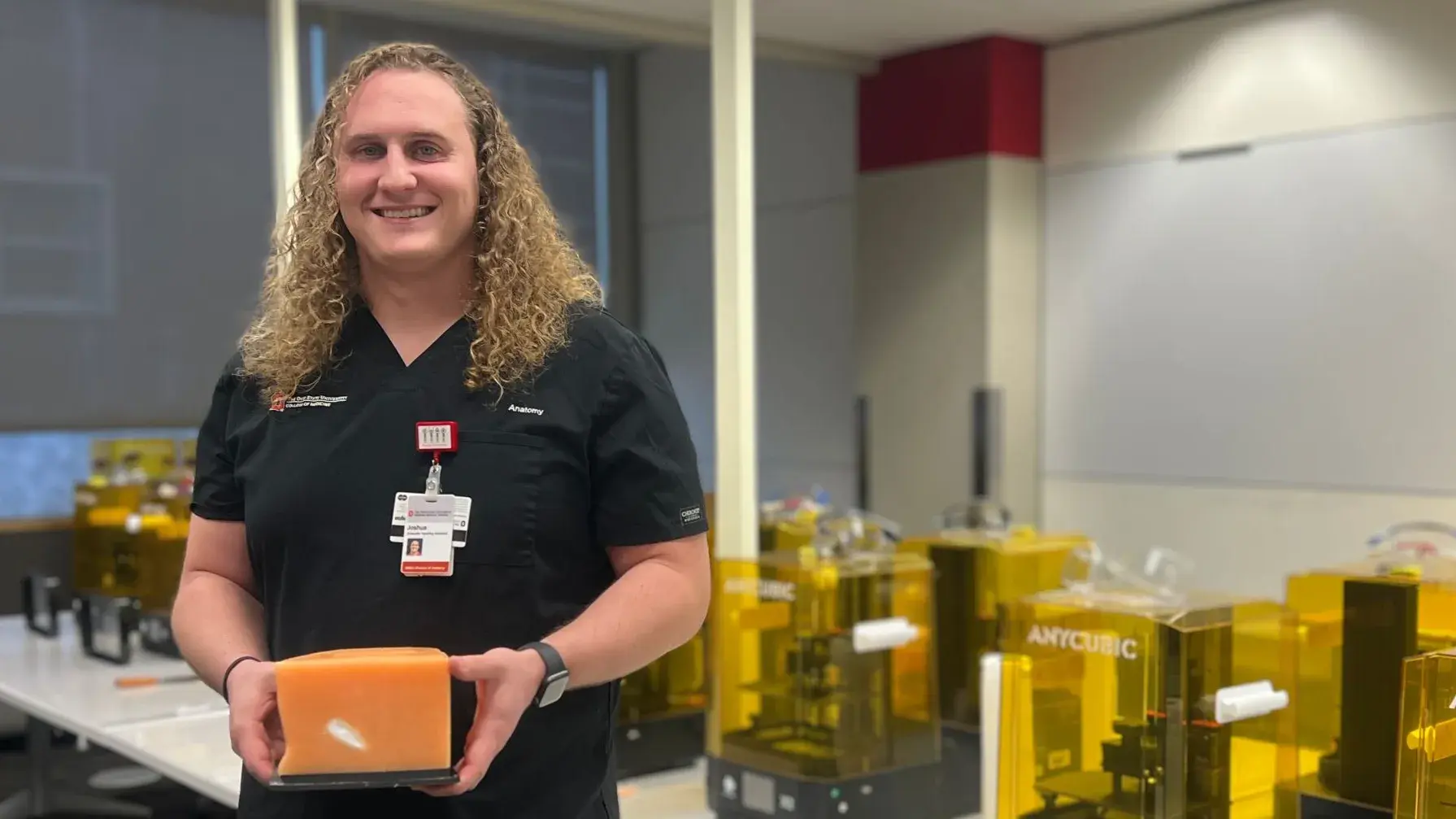 Joshua Krech standing and smiling in front of a row of 3D printers, holding a 3D printed cube.