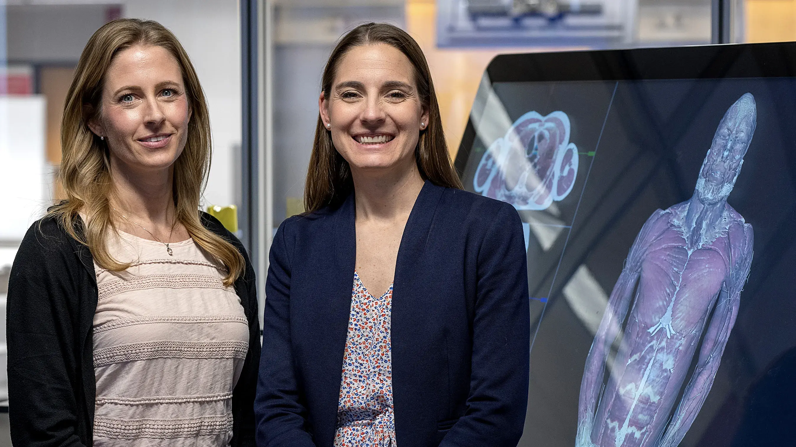 Kristen Roberts and Laura Boucher standing and smiling in front of a large digital interactive display of human anatomy.
