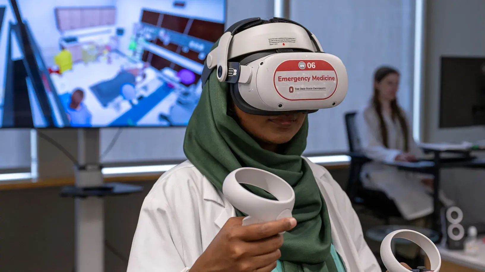 Female student in lab coat wearing a virtual reality head set and holding controllers, engaging with a hospital scenario on the TV behind her.