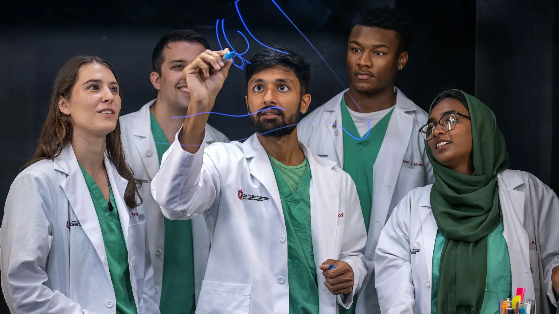 Five students in lab coats and scrubs, smiling while one student writes on a glass see-through board.