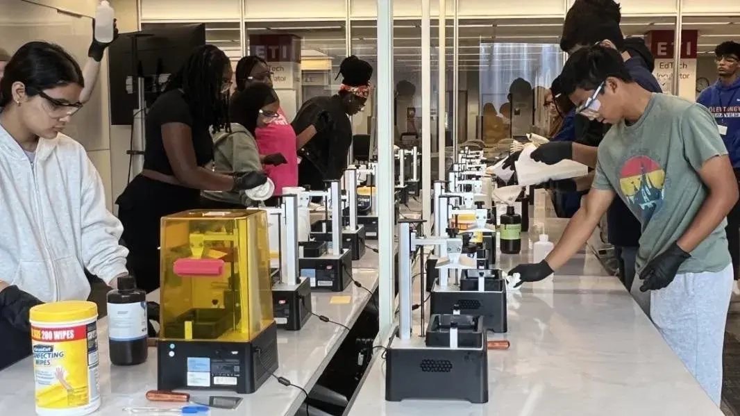 Students surrounding the 3D Printing Lab stations, wearing safety goggles while interacting with the equipment.